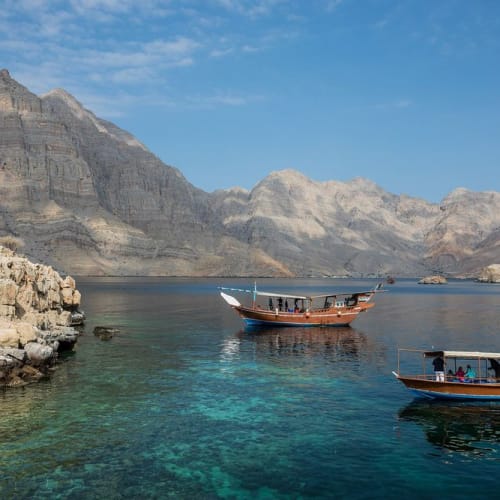 Zwei traditionelle Holzboote schwimmen in den klaren Wasser des Musandam-Halbinsels mit zerklüfteten Bergen im Hintergrund.