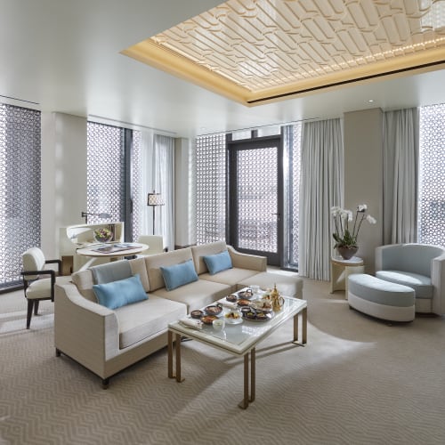 Luxuriöse Hotel-Suite mit weißen Sofas, großen Fenstern mit gemusterten Vorhängen und eleganter Einrichtung.