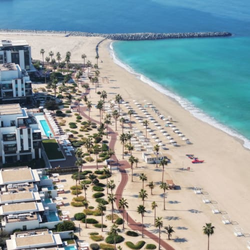 Aerial view of Nikki Beach Resort and Spa Dubai showing a beach with sun loungers, palm trees, and modern buildings nearby.