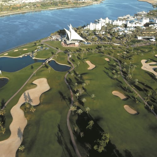 Luftaufnahme eines grünen Golfplatzes mit Bunkern, Wasserflächen und einem modernen Gebäude in der Nähe eines Flusses.