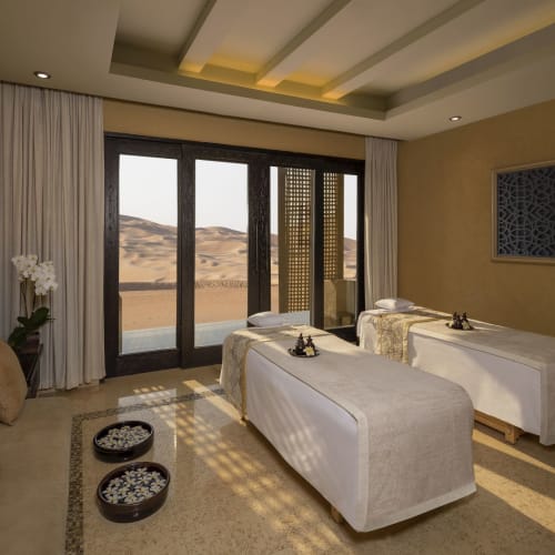 Luxuriöses Spa-Zimmer mit zwei Massagebänken, großen Fenstern mit Wüstenblick und eleganter Dekoration.