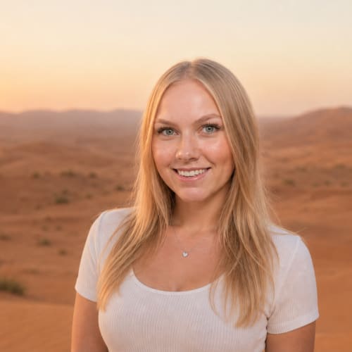 Young woman with long blonde hair and a white top smiling in a desert landscape at sunset with mountains in the background.