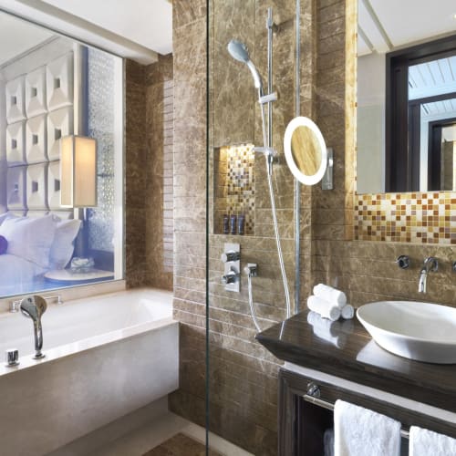 Luxurious hotel bathroom with a large bathtub, a walk-in shower with a mirror, and a vanity with a sink, overlooking a well-lit hotel room.