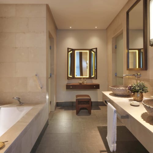 Luxuriöses Badezimmer mit beigen Stewwänden, Doppelwaschbecken mit Steinbeckenschalen und einer großen Badewanne, beleuchtet durch warmes Licht und eine dekorative Wandlampe.