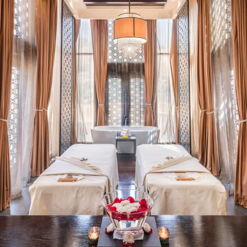 Spa treatment area with two massage beds in a luxurious room decorated with patterned walls, gold curtains, and a small fountain in the background.