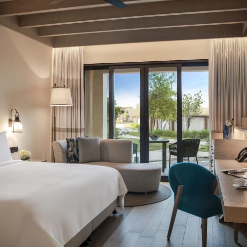 A modern hotel room with a large bed, a sitting area, and a view of a garden through glass sliding doors.