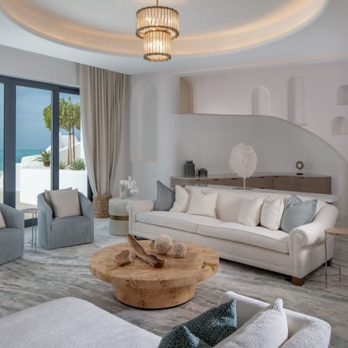 Luxuriöses Wohnzimmer mit weißen und grauen Möbeln, einem hölzernen Couchtisch und einer Schiebetür mit Blick auf den Strand.