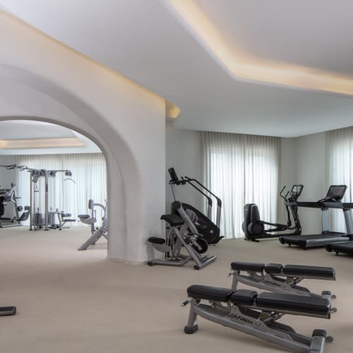 Moderater Fitnessraum mit verschiedenen Trainingsgeräten und großen Fenstern mittransparenten Vorhängen in einem geräumigen Raum.