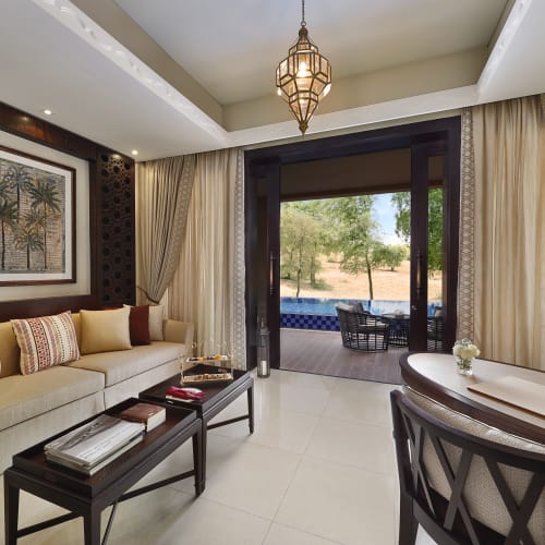 Elegant living room with large sliding glass doors opening to a desert landscape and pool outside, decorated with a beige sofa, a contemporary chandelier, and luxurious curtains.