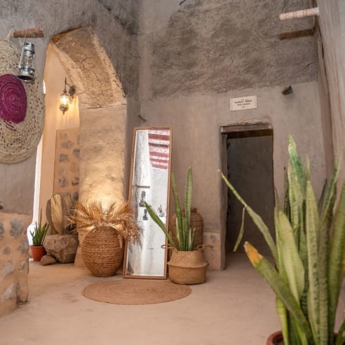 Innenflur mit rustikalen Steinwänden, einer großen gewebten Mütze an der Wand, einem hohen Spiegel, mehreren gewebten Körben, Topf-Schlangenpflanzen und warmem, weichem Licht.
