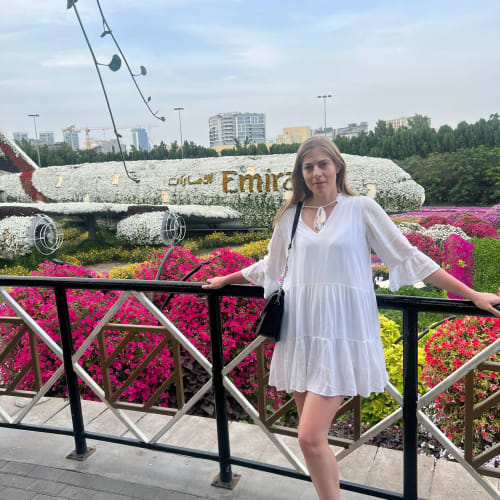 Eine junge Frau in einem weißen Kleid steht an einem schwarzen Geländer vor einem Blumenbeet und einer blumengeschmückten Hubschrauber-Installation mit der Aufschrift 'Emirates' in einem Stadtpark.