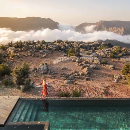 Eine Frau in einem roten Kleid steht am Rand eines Pools mit Blick auf eine felsige und grüne Hügellandschaft mit Bergen und Wolken im Hintergrund bei Sonnenaufgang oder Sonnenuntergang.