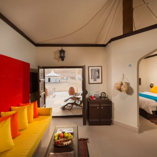 Innenansicht eines Wüstencamps mit einem Sitzbereich mit gelben Kissen und einem Bett mit bunten Kissen im angrenzenden Zimmer.