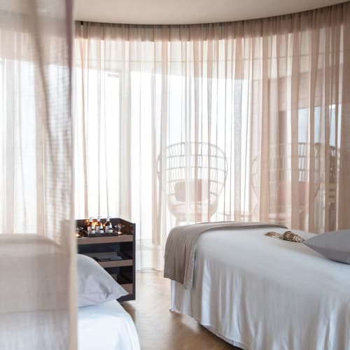 Luxuriöses Hotelzimmer mit zwei Betten und großen Fenster mit Vorhängen, Blick auf einen Balkon mithängenden Stühlen.