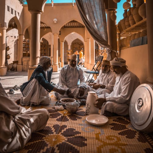 Gruppe von Männern in traditioneller nahöstlicher Kleidung, die auf einem gemusterten Teppich in einem Gebäude mit gewölbten Türöffnungen sitzen und in einer warmen, sozialen Umgebung Essen und Trinken teilen.