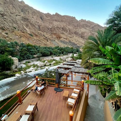 Überdachte Terrasse mit Holzstühlen und Tischen mit Blick auf einen Fluss, umgeben von grünen Palmen und felsigen Bergen unter klarem blauem Himmel.