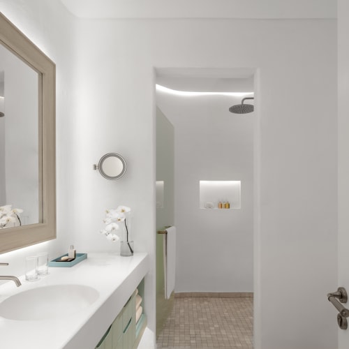 A modern, minimalist bathroom with a white vanity and large mirror, connected to a shower area with a ceiling rain shower head.