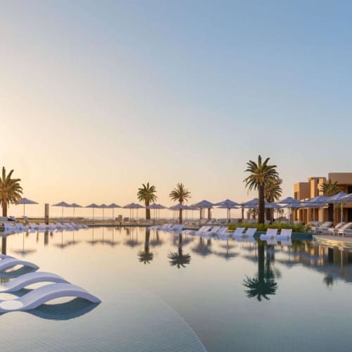 A luxurious hotel pool area at sunset with lounge chairs, umbrellas, and palm trees reflecting in the water.