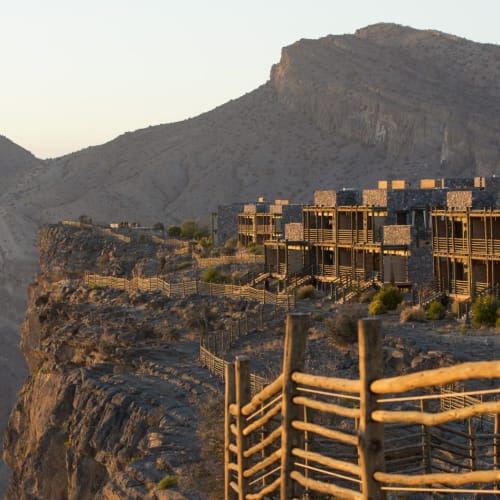 Modern hotel complex built along a rocky mountain ridge in a rugged landscape, with a wooden fence in the foreground.