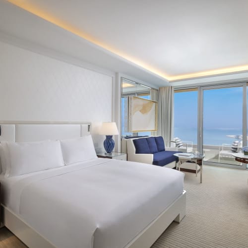 Luxurious hotel room with large bed, nightstands with lamps, armchair, sofa, and large window with sea view