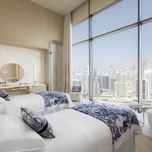 Elegant hotel room with two neatly made beds, large floor-to-ceiling windows overlooking a city skyline, and modern decor.