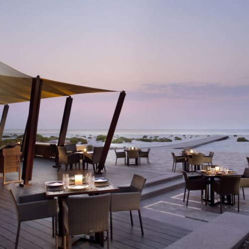 Ein Außenessbereich bei Sonnenuntergang auf einer Terrasse mit Blick auf das Meer, mit gedeckten Tischen und Sonnenschirmen.