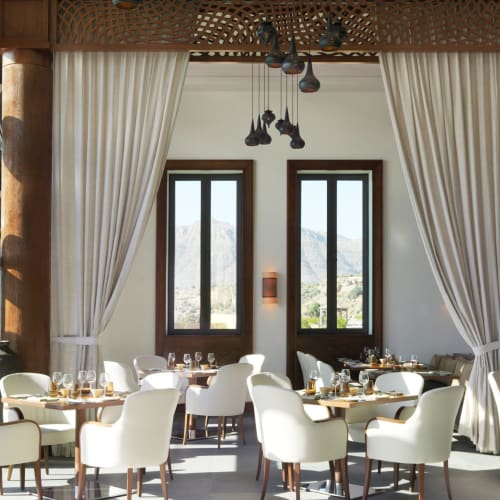 Eleganter Speisesaal mit weißen Stühlen, Tischen, die für Mahlzeiten gedeckt sind, großen Fenstern und cremefarbenen Vorhängen im Anantara Al Jabal Al Akhdar Resort in Oman.