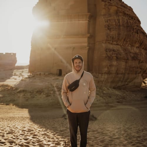 A man standing on sand with ancient ruins in the background during sunset.