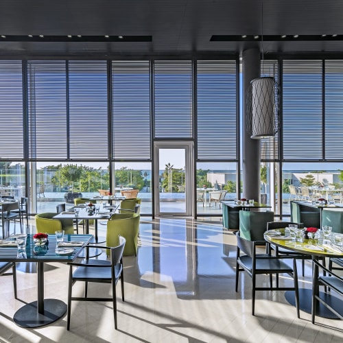Elegant dining area inside The Oberoi Beach Resort Al Zorah with stylish chairs and large windows with window blinds.