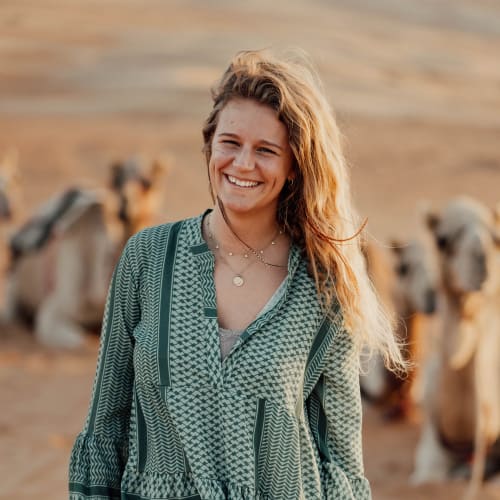A smiling young woman with wavy hair stands outdoors with camels in the background.