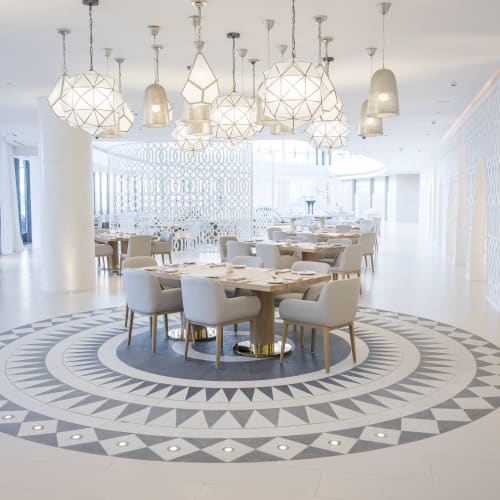 Eleganter, heller Speiseraum mit weißen Stühlen um einen runden Tisch, hängende Leuchten darüber und ein gemustertes Bodendesign.