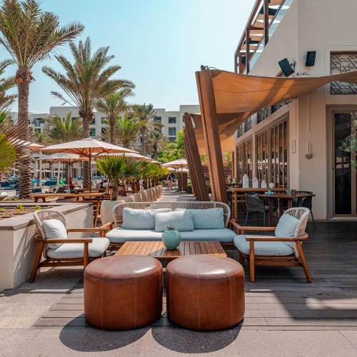 A stylish outdoor seating area with sofas, armchairs, and poufs on a terrace with palm trees and umbrellas in the background.
