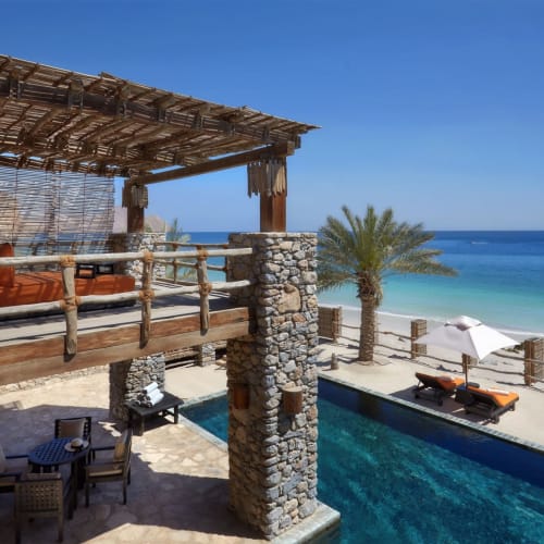 A luxurious beachfront resort with a pool, seating area, and a balcony overlooking a sandy beach and the ocean.
