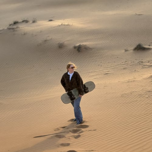 Eine Frau hält ein Sandboard und steht auf Sanddünen bei Sonnenuntergang, hinterlässt Fußspuren.