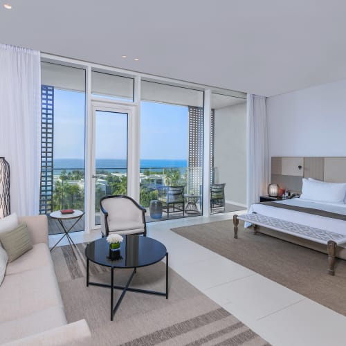 A spacious hotel room with a large bed, a beige sofa, and a balcony with ocean view.