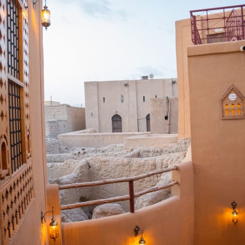 Blick auf einen traditionellen Nahost-Hof mit beigen adobeartigen Wänden, dekorativen Laternen und einem Blick auf antike Steinstrukturen und Gebäude im Hintergrund unter bewölktem Himmel.