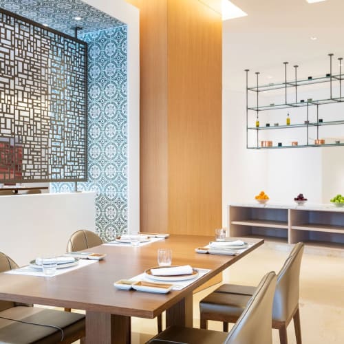 A modern dining area in a wellness resort with a wooden table set for four, surrounded by comfortable chairs and decorative shelving in the background.