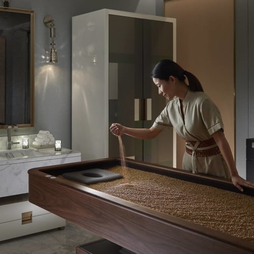 Eine Frau in Spa-Kleidung, die sich um einen großen Tisch mit Partikeln kümmert, möglicherweise Körner oder Kräuter, in einem luxuriösen, sanft beleuchteten Raum.