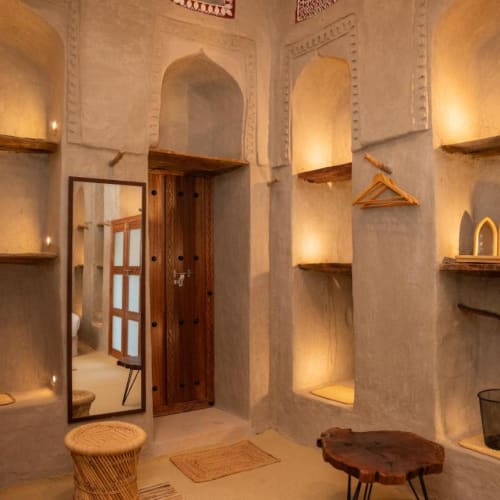 Innenraum eines traditionell nahöstlich gestalteten Zimmers mit eingebauten Nischenregalen, die von warmem Licht beleuchtet werden, einer Holztür, einem stehenden Spiegel, einem geflochtenen Hocker und einem kleinen Holztisch.