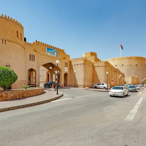 Blick auf das historische Nizwa Fort in Oman mit beigen Steinmauern, parkenden und fahrenden Autos in der Nähe und klarem blauem Himmel.