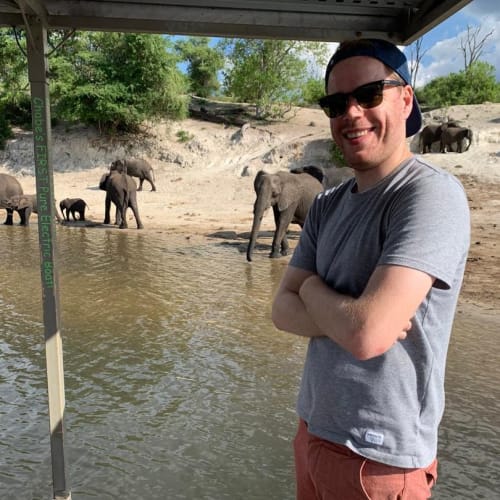 Ein lächelnder Mann mit Sonnenbrille, grauem T-Shirt und einer Cap, steht mit verschränkten Armen in der Nähe eines Flusses, im Hintergrund trinken und laufen Elefanten.