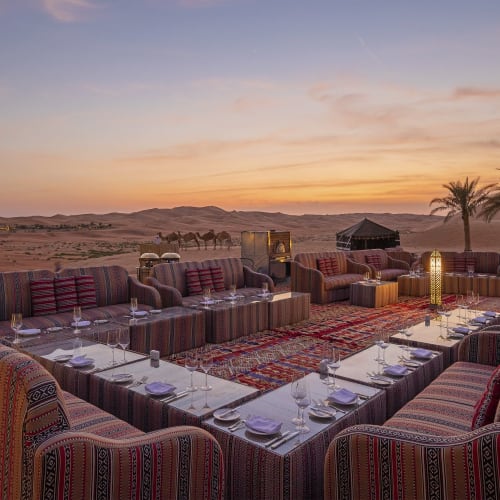 Eine luxuriöse Outdoor-Essens-Setup mit gepolsterten Sitzgelegenheiten in U-Form, Blick auf eine Wüstenlandschaft bei Sonnenuntergang, mit Laternen und Palmen im Hintergrund.