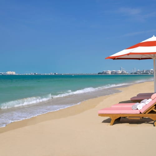 Beach chairs and an umbrella on a sandy beach with clear blue water and a city skyline in the background.