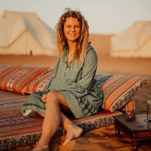 A smiling woman sitting on a colorful cushioned mattress on the desert sand during sunset in Oman.