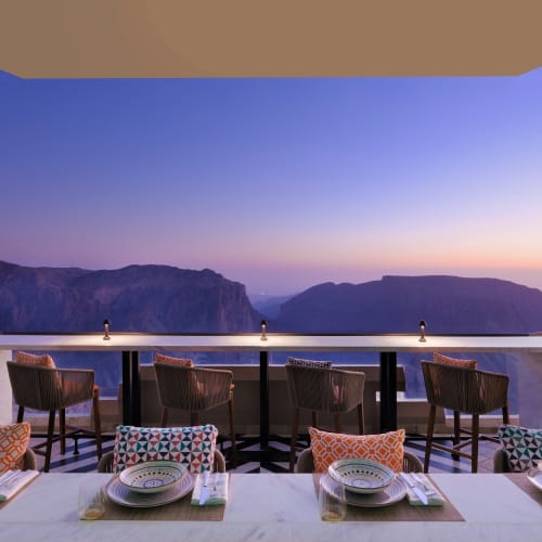 Blick vom Dachterrassensalon im Hotel Indigo Jabal Akhdar Resort & Spa mit langem gedecktem Esstisch, der auf Berge bei Sonnenuntergang blickt.