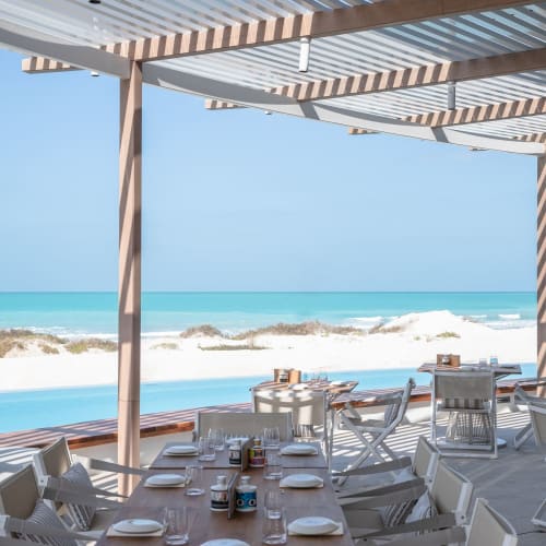 Outdoor dining area on a beachside terrace with tables set for a meal, overlooking the ocean with sandy beach and waves.