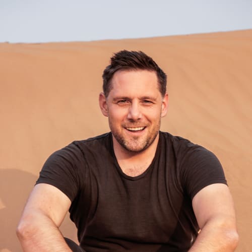 A smiling man with short dark hair and a beard sitting outdoors against a desert landscape.