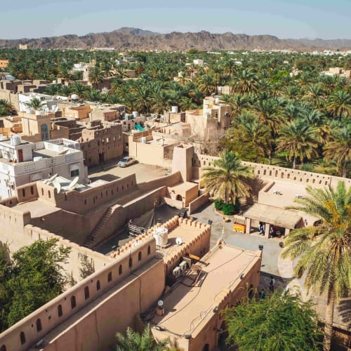 Luftaufnahme einer traditionellen Festung mit Befestigungswänden, umgeben von grünen Palmen und einer modernen Stadt in Oman.