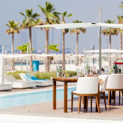 Ein luxuriöser Außenbereich zum Essen im Nikki Beach Resort & Spa Dubai neben einem Pool mit Liegestühlen und Sonnenschirmen, mit Blick auf Palmen.