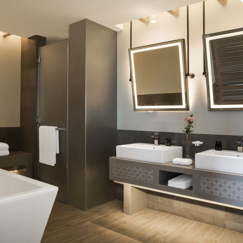 Modern bathroom with two square vessel sinks, illuminated mirrors, and a freestanding bathtub in an elegant hotel setting.